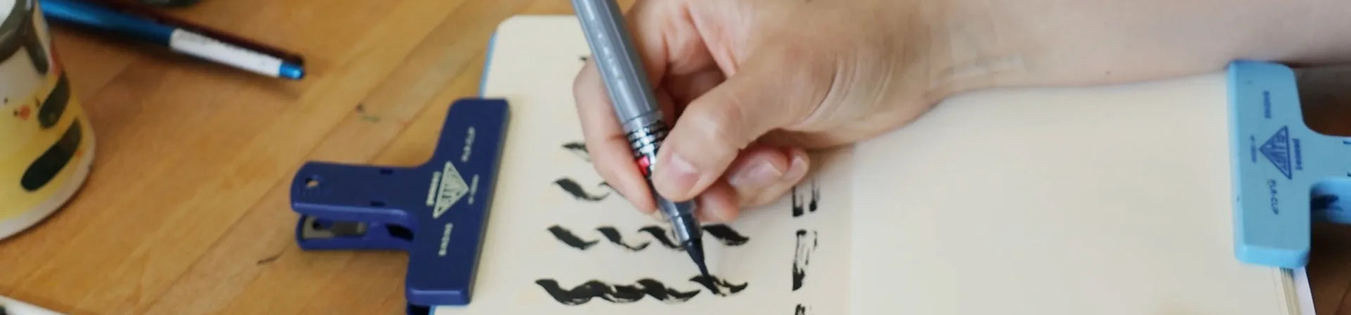 Person practicing calligraphy on a notebook with a pen and ruler on a wooden table.